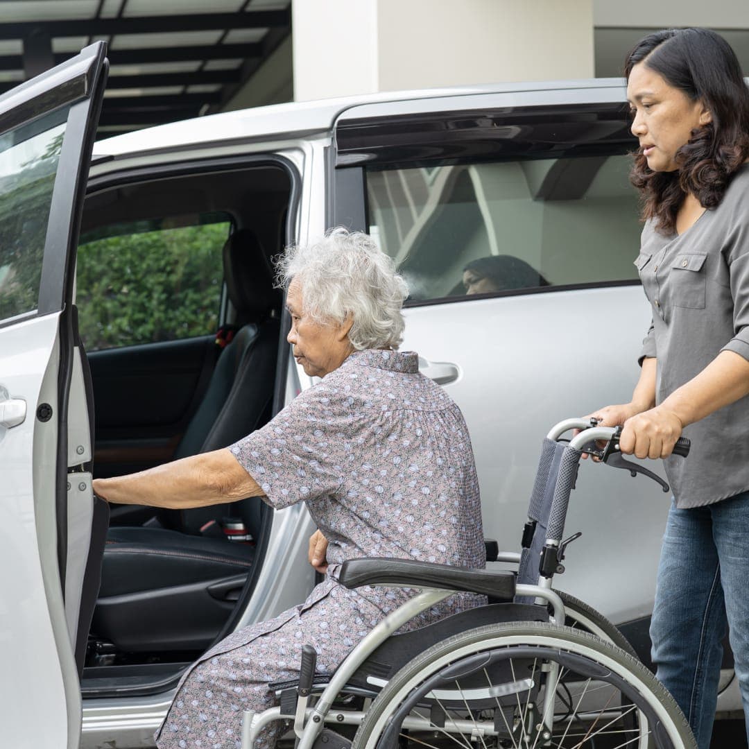 A carer holding a wheelchair while an older woman moves into the car
