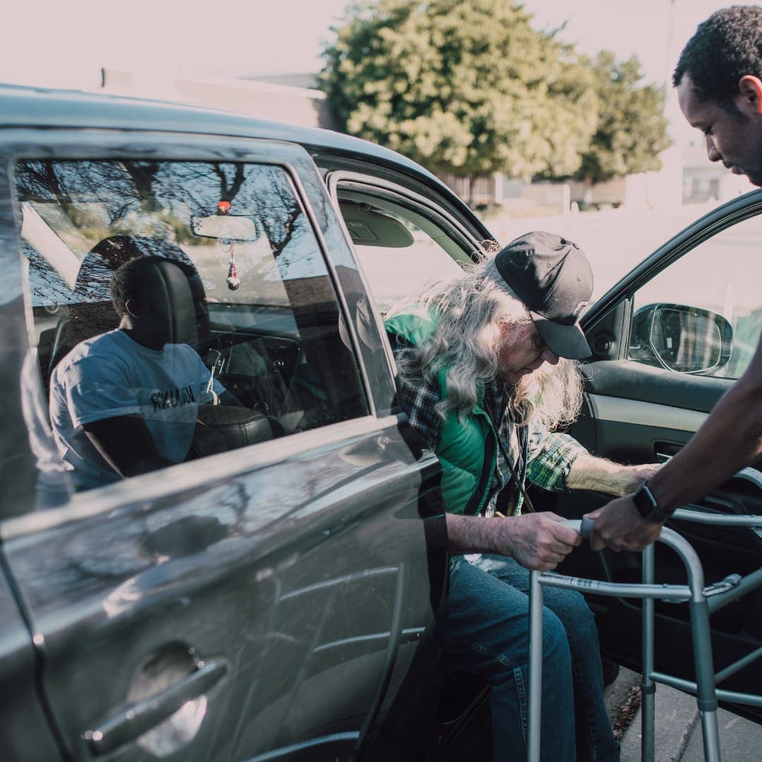 A driver steadying a passenger with a walker as he gets out of the car